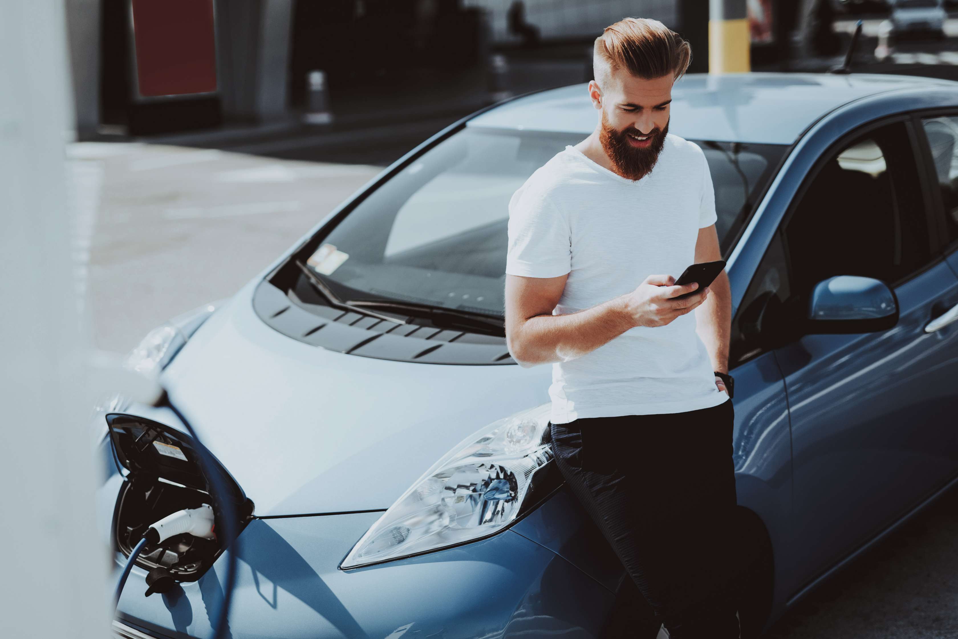 Mann mit Handy lehnt an E-Auto, das geladen wird
