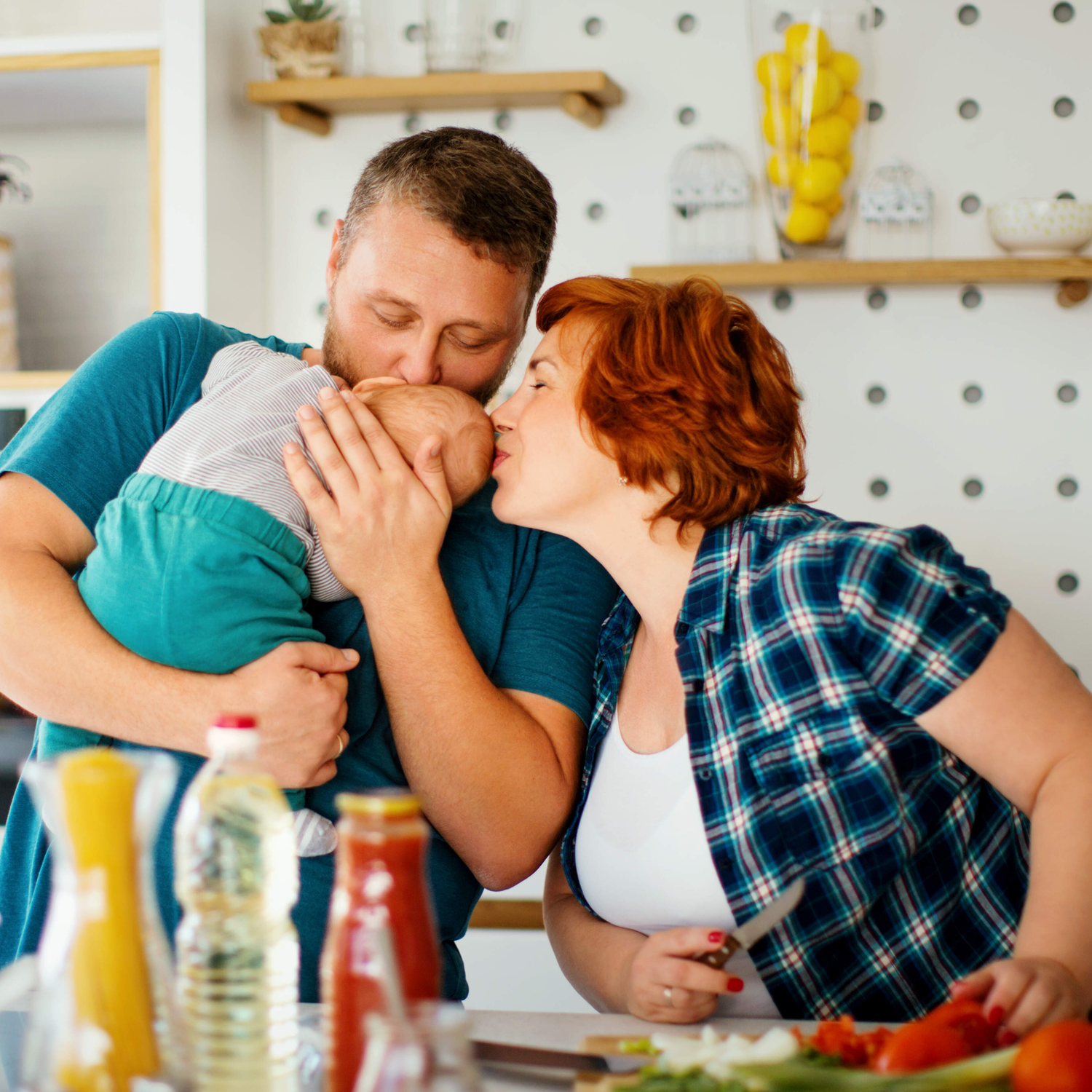 Seitenansicht einer Familie bei der Zubereitung einer gemeinsamen Mahlzeit. Ihr Baby schläft in Papas Armen und beide Eltern küssen es vor Freude.