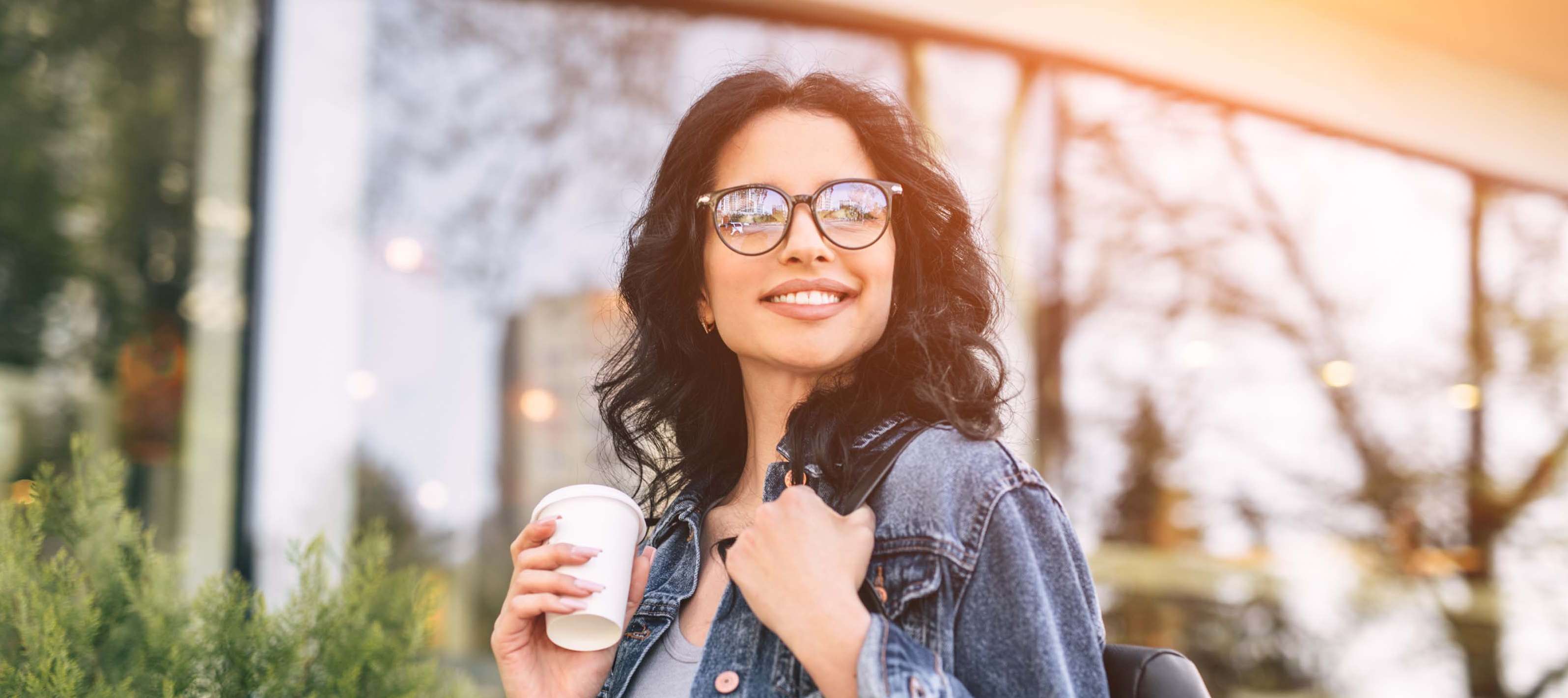 Junges Mädchen mit Brille steht lächelnd auf einer Straße mit einem Kaffee in der Hand