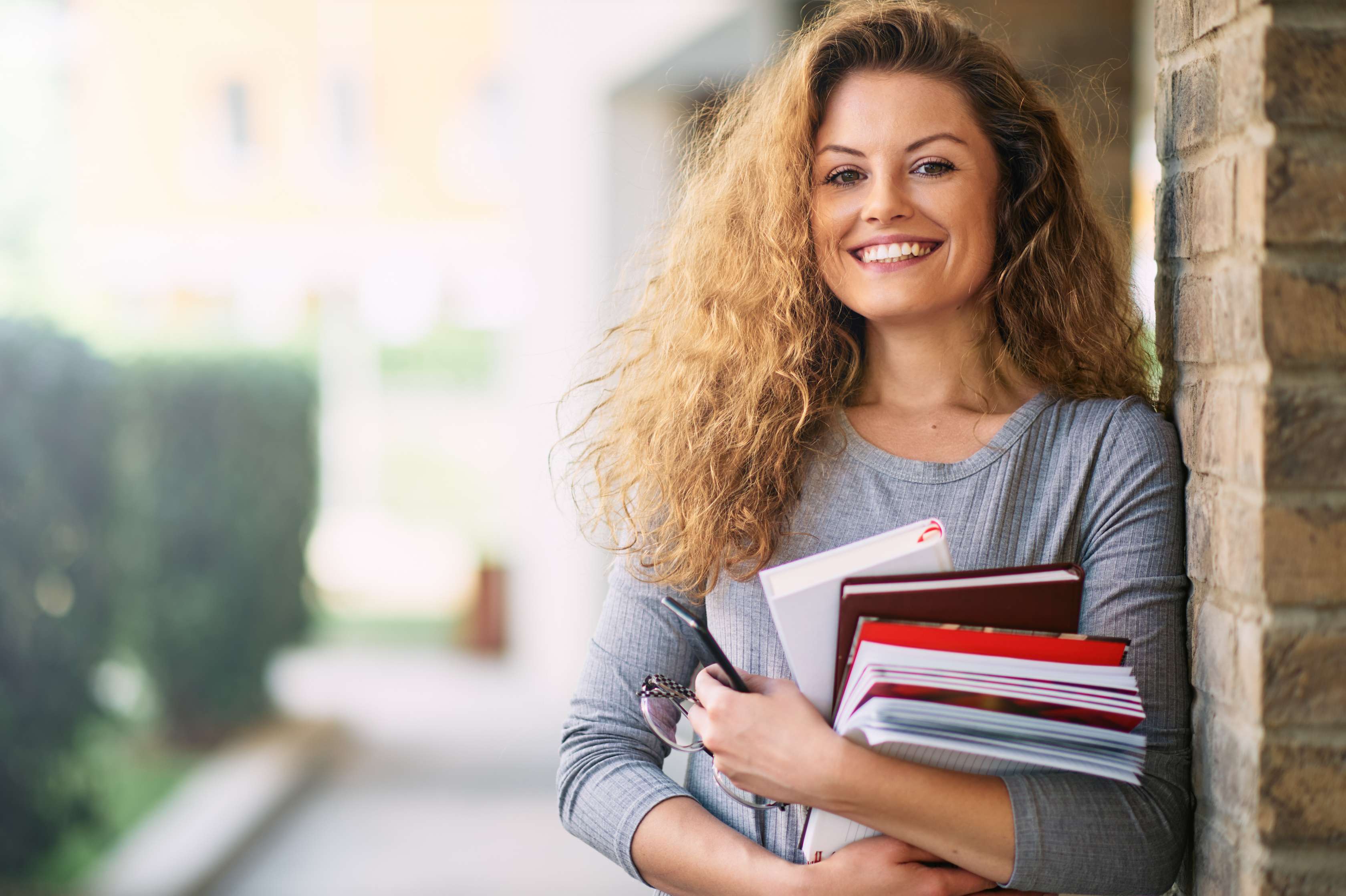 Junge Lehrerin lehnt an einer Wand, hat Bücher in der Hand und lächelt