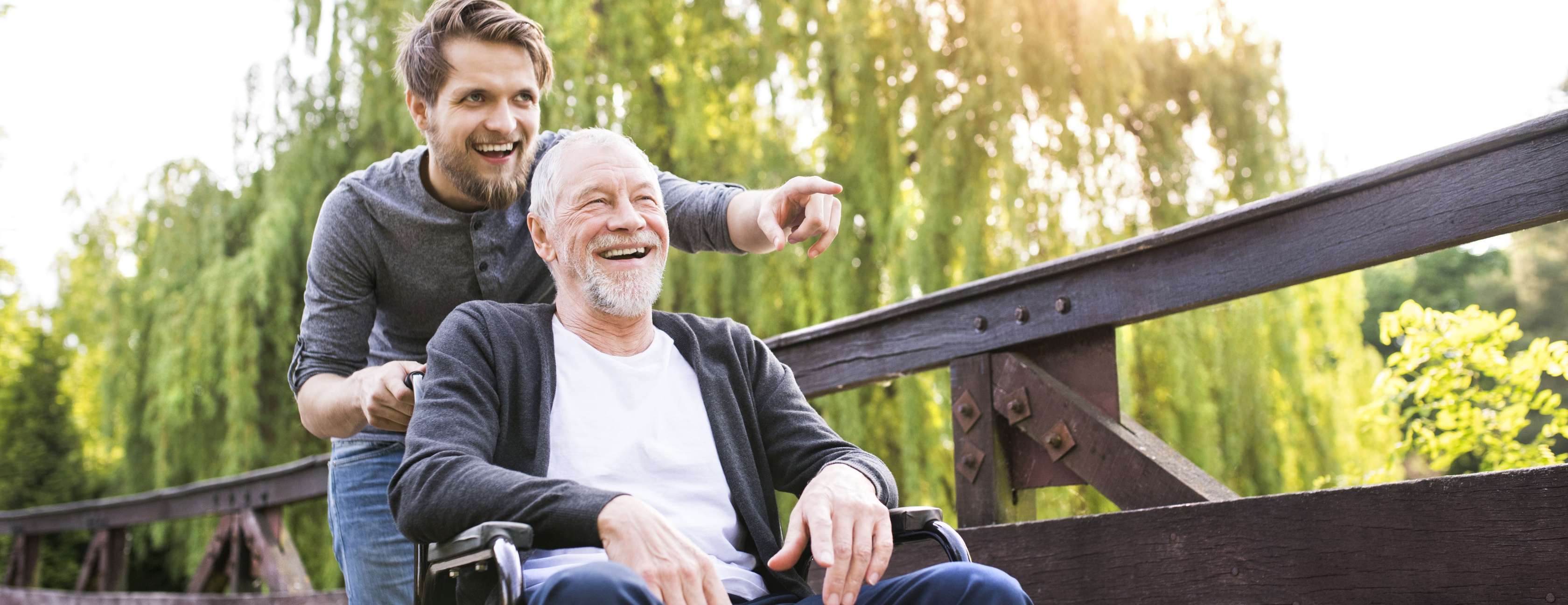 Großvater sitzt im Rollstuhl und junger Mann schiebt ihn über eine Brücke durch den Park