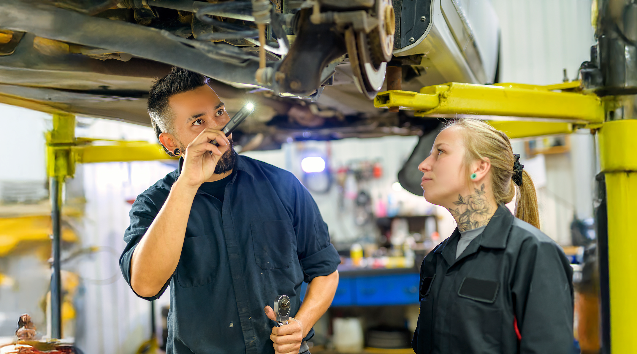 Kfz-Mechatroniker und -Mechatronikerin unter Hebebühne betrachten Autokarosserie