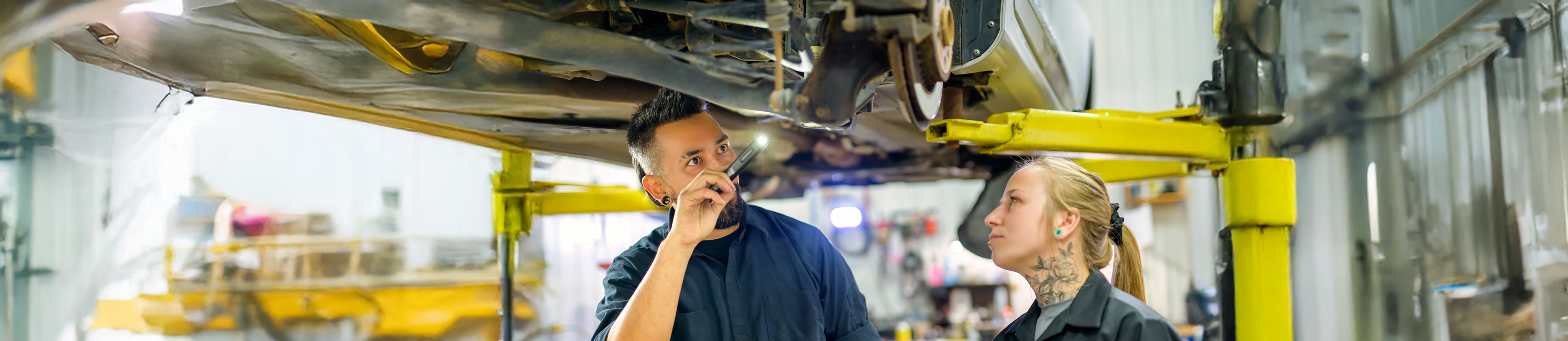 Kfz-Mechatroniker und -Mechatronikerin unter Hebebühne betrachten Autokarosserie